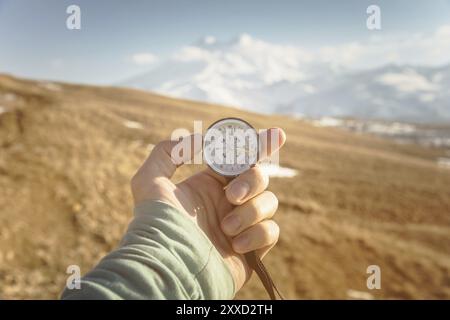 La main d'un homme tient une boussole à main sur fond de montagnes et de collines au coucher du soleil. Le concept de voyage et de navigation dans les espaces ouverts Banque D'Images