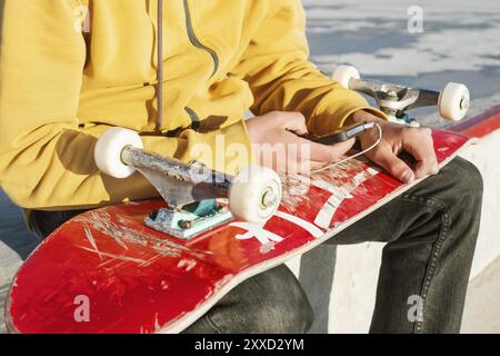 Gros plan d'un adolescent vêtu d'un sweat-shirt avec un Jean et des baskets assis dans un skate Park et tenant un téléphone avec des écouteurs et une planche à roulettes Banque D'Images