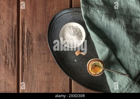 Un oeuf dur dans une tasse de gros sel de mer et une cuillère, tourné par le haut sur un fond de bois rustique foncé avec une place pour le texte Banque D'Images