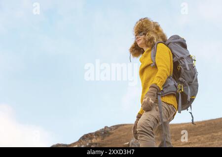 La fille de routard dans des lunettes de soleil et un grand chapeau de fourrure nordique avec un sac à dos sur son dos se tient sur un rocher et regarde vers les falaises se cachant dedans Banque D'Images