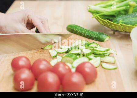 Gros plan des mains femelles coupées en concombres frais coupés sur une planche à découper en bois à côté des tomates roses. Le concept de cuisine végétarienne maison et Banque D'Images