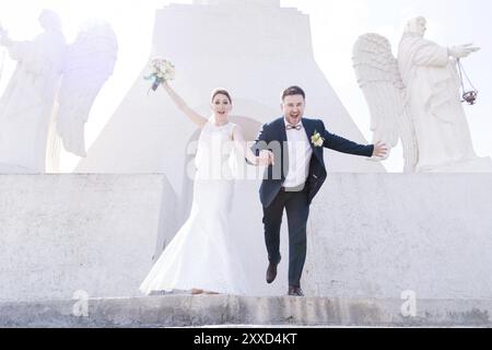 Portrait d'un beau couple de jeunes mariés un jour de mariage avec un bouquet dans les mains courant dans les escaliers riant et souriant sur fond Banque D'Images