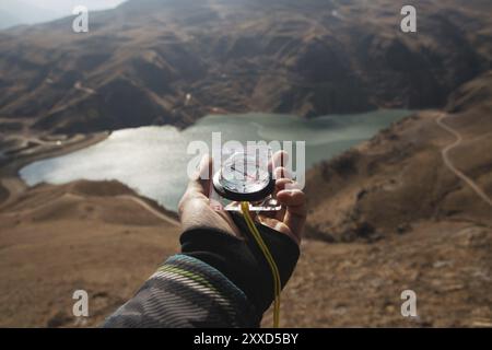 La main d'un homme tient une boussole magnétique de poche pour la navigation sur la toile de fond d'une pente rocheuse et d'un lac de montagne. Le concept de trouver un moyen. G Banque D'Images