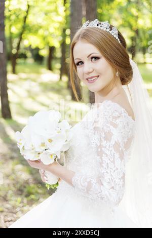 Portrait d'une belle mariée dans une grande et luxuriante robe blanche avec un bouquet de fleurs dans les mains au milieu des rayons chauds du soleil dans une forêt verdoyante Banque D'Images