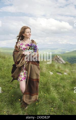 Belle fille blonde enveloppée dans un plaid stands avec un bouquet de fleurs sauvages et regarde le regard sur le fond d'un beau paysage naturel Banque D'Images