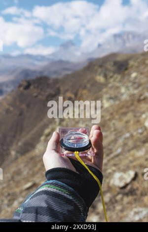 La main d'un homme tient une boussole magnétique de poche pour la navigation sur le fond d'une pente rocheuse et des rochers épiques sous un ciel bleu et des nuages blancs. Banque D'Images