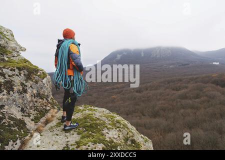 Un alpiniste professionnel dans une veste en duvet rouge avec une baie de corde bleue se tient sur un haut rocher et contemplant la vue au loin. Le concept Banque D'Images