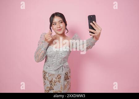 Mignonne femme indonésienne en kebaya moderne posant pour un selfie tout en faisant un geste de coeur de doigt, isolé par fond rose. Banque D'Images