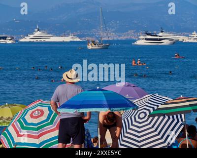 Scène de plage avec touriste en chapeau de paille regardant les yachts et superyachts ancrés dans la baie au large d'Antibes, Côte d'Azur, France. Banque D'Images