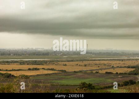 Hadleigh Castle, Royaume-Uni. 24 août 2024. Un paysage en couches montrant des champs agricoles verts et bruns au premier plan, séparés par un canal ou une rivière d'un complexe industriel en arrière-plan. Une brume épaisse ou un brouillard obscurcit les bâtiments éloignés, suggérant une pollution ou une humidité atmosphérique. Le ciel couvert ajoute un ton sombre, soulignant la tension environnementale entre l'utilisation des terres rurales et le développement urbain. Commencez votre week-end de vacances au château de Hadleigh, surplombant l'estuaire de la Tamise. Penelope Barritt/Alamy Live News Banque D'Images