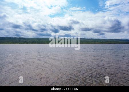 Un lac serein avec des eaux claires s'étend vers des montagnes lointaines sous un ciel bleu lumineux avec des nuages dispersés, créant un cadre paisible et image Banque D'Images