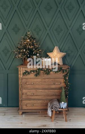Commode en bois décorée pour Noël avec guirlande, petit arbre de Noël, étoile illuminée et cadeau enveloppé contre le mur à motifs verts Banque D'Images