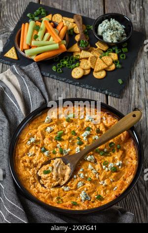 sauce au poulet buffalo dans un plat de cuisson sur une table en bois rustique gris avec des craquelins, du fromage bleu, des bâtonnets de carotte et de céleri sur une planche d'ardoise en arrière-plan, vert Banque D'Images