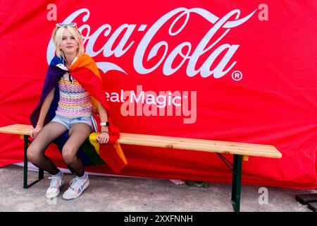 Une fille avec un drapeau aux couleurs LGBTQ assis sur un banc avec le logo Caca Cola derrière elle, adolescente, adolescent, été, adolescent Banque D'Images