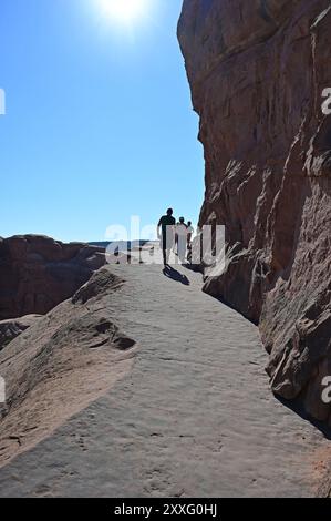 Randonneurs rétro-éclairés sur la section exposée de Delicate Arch Trail dans le parc national d'Arches près de Moab, Utah, par un matin d'été ensoleillé et clair. Banque D'Images