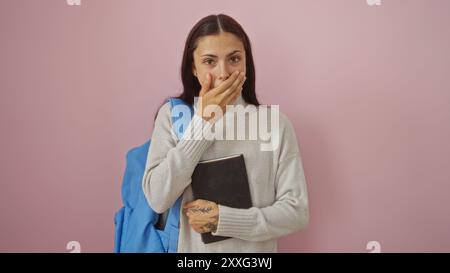 Jeune femme brune avec un tatouage couvrant sa bouche tenant un livre sur un fond rose avec une expression surprise Banque D'Images