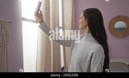Jeune femme prenant une photo avec son téléphone dans un cadre intérieur avec une toile de fond de rideaux et de suspensions murales. Banque D'Images