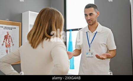 Un thérapeute masculin parle à une patiente dans une clinique de réadaptation avec un tableau du système musculaire en arrière-plan. Banque D'Images