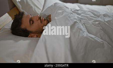 Un jeune homme hispanique dort paisiblement dans une chambre avec des draps blancs, transmettant un sentiment de repos et de détente dans un cadre résidentiel. Banque D'Images