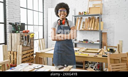 Femme confiante avec des lunettes de sécurité et des écouteurs se tient les bras croisés dans un atelier de menuiserie lumineux. Banque D'Images