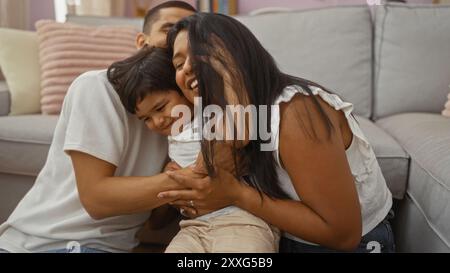 Famille hispanique profitant d'un moment d'amour ensemble à la maison dans un salon confortable où un homme, une femme et un enfant partagent des étreintes joyeuses sur le sol dans un wa Banque D'Images