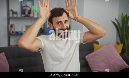 Un homme d'âge mûr ludique avec une barbe fait des oreilles de lapin drôles avec ses mains dans un salon moderne. Banque D'Images