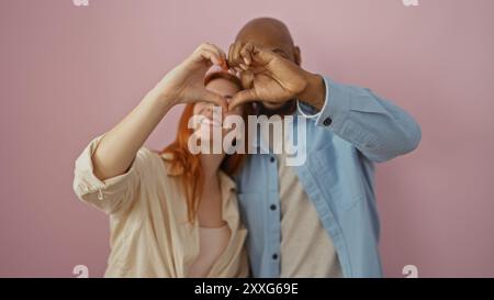 Couple interracial montrant l'amour et le bonheur en faisant une forme de coeur avec leurs mains sur un fond rose. Banque D'Images