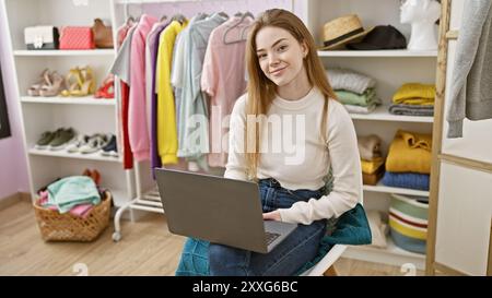 Une jeune femme blonde est assise dans un dressing bien organisé avec divers vêtements, travaillant sur son ordinateur portable. Banque D'Images