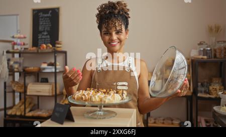 Jeune femme aux cheveux bouclés tenant un plat de service dans une boulangerie entourée de pâtisseries et souriante tout en portant un tablier Banque D'Images