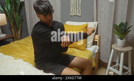 Jeune homme hispanique dans une chambre est assis sur un lit ajustant sa chemise noire, entouré d'un décor intérieur confortable comprenant des oreillers, des plantes et un éclairage chaleureux. Banque D'Images