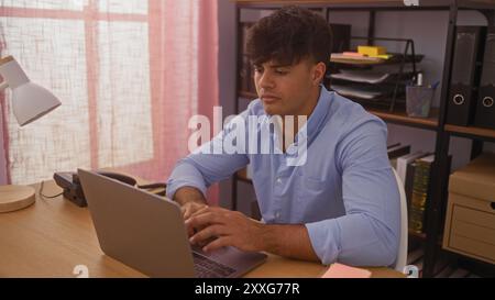 Jeune homme hispanique travaillant sur un ordinateur portable dans un cadre de bureau avec des étagères en arrière-plan affichant diverses fournitures de bureau. Banque D'Images