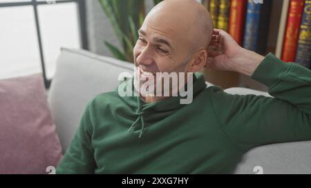 Souriant homme hispanique chauve avec barbe portant un sweat à capuche vert se relaxant à l'intérieur sur un canapé Banque D'Images