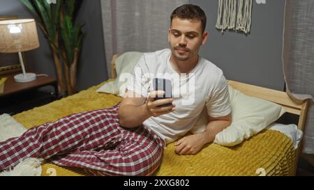 Handsone jeune homme hispanique avec une barbe se prélassant sur son lit dans une chambre confortable, en utilisant son smartphone dans un cadre intérieur. Banque D'Images