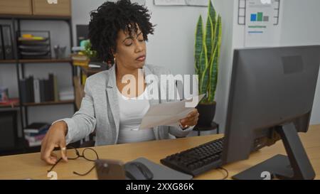 Une belle jeune femme afro-américaine aux cheveux bouclés travaille dans un bureau, examinant un document tout en tenant ses lunettes, représenté dans un moderne Banque D'Images