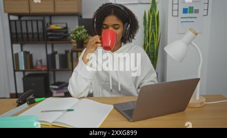 Jeune femme afro-américaine aux cheveux bouclés travaillant dans un bureau, sirotant du café dans une tasse tout en utilisant un ordinateur portable, entourée de livres et de soutiens de bureau Banque D'Images