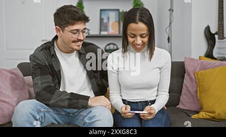 Un couple souriant assis sur un canapé dans leur maison, heureux de regarder un résultat de test de grossesse. Banque D'Images