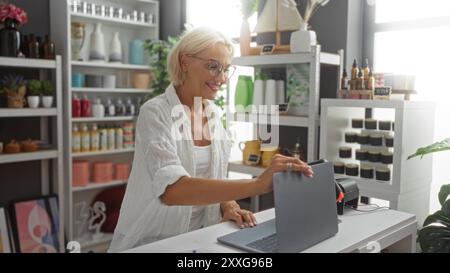 Femme souriante tout en travaillant avec un ordinateur portable dans le magasin de décoration à la maison, entourée de diverses décorations et plantes colorées Banque D'Images