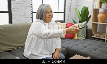 Une femme âgée assise confortablement sur un canapé à la maison, tenant une télécommande avec plante et étagère en arrière-plan. Banque D'Images