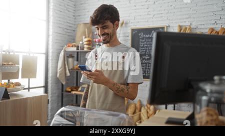 Jeune homme hispanique avec une barbe souriant tout en utilisant un smartphone dans un intérieur de boulangerie avec des étagères de pain et de pâtisseries autour Banque D'Images