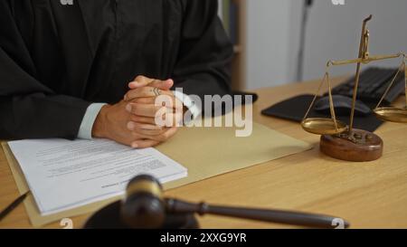 Jeune homme dans la robe d'un juge avec les mains attachées à un bureau dans un cadre de bureau, avec des documents juridiques, marteau, et écailles de justice visibles sur la bois Banque D'Images