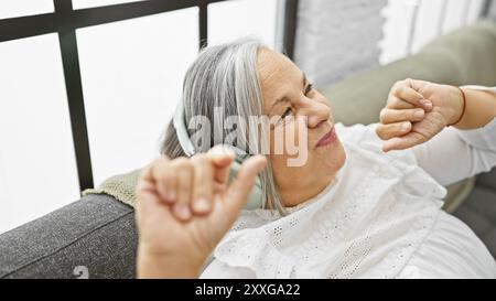 Une femme âgée aime la musique avec des écouteurs, souriant dans un salon à l'atmosphère chaleureuse. Banque D'Images
