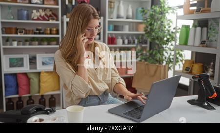 Jeune femme blonde travaillant sur ordinateur portable et parlant au téléphone dans un magasin de décoration entourée de divers objets décoratifs Banque D'Images