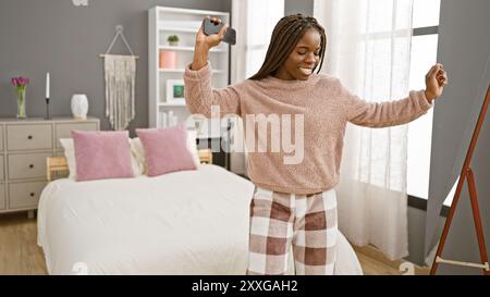 Une femme afro-américaine avec des tresses danse joyeusement dans sa chambre, tenant un smartphone et portant un pyjama Banque D'Images