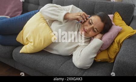 Une femme hispanique d'âge moyen se détend sur un canapé à la maison, parlant au téléphone avec une expression calme. Banque D'Images