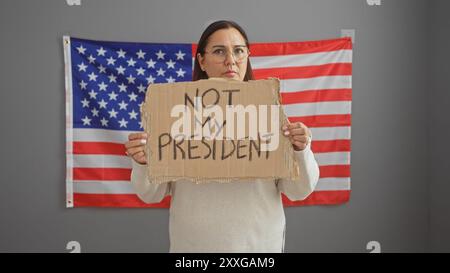 Femme hispanique d'âge moyen tenant une pancarte de protestation devant le drapeau américain dans un bureau Banque D'Images