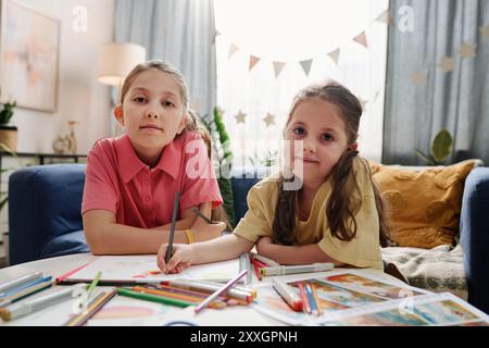 Deux jeunes filles assises à la table ronde, à colorier avec des crayons. Chambre lumineuse, plantes et décorations étoilées en arrière-plan, créant une atmosphère joyeuse Banque D'Images