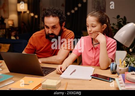 Homme barbu aidant la jeune fille avec les devoirs, tous deux concentrés sur l'écran d'ordinateur portable, divers articles de papeterie sur le bureau autour d'eux. Père fournissant de l'aide à la fille dans un environnement familial confortable Banque D'Images