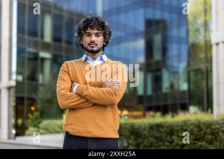 Jeune homme confiant avec les cheveux bouclés debout avec les bras croisés devant le bâtiment de verre moderne. Cadre urbain suggérant une atmosphère professionnelle et un style de vie moderne. Banque D'Images