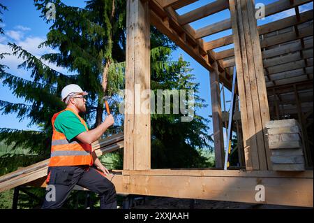 Menuisier construisant une maison à ossature en bois près de la forêt. Homme barbu martelant des clous dans la structure tout en portant un casque de protection et un gilet de construction. Concept de construction écologique moderne. Banque D'Images