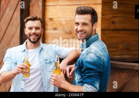 Réunion de vieux amis. Deux jeunes hommes joyeux tenant des bouteilles de bière et regardant la caméra tout en se penchant au comptoir du bar Banque D'Images